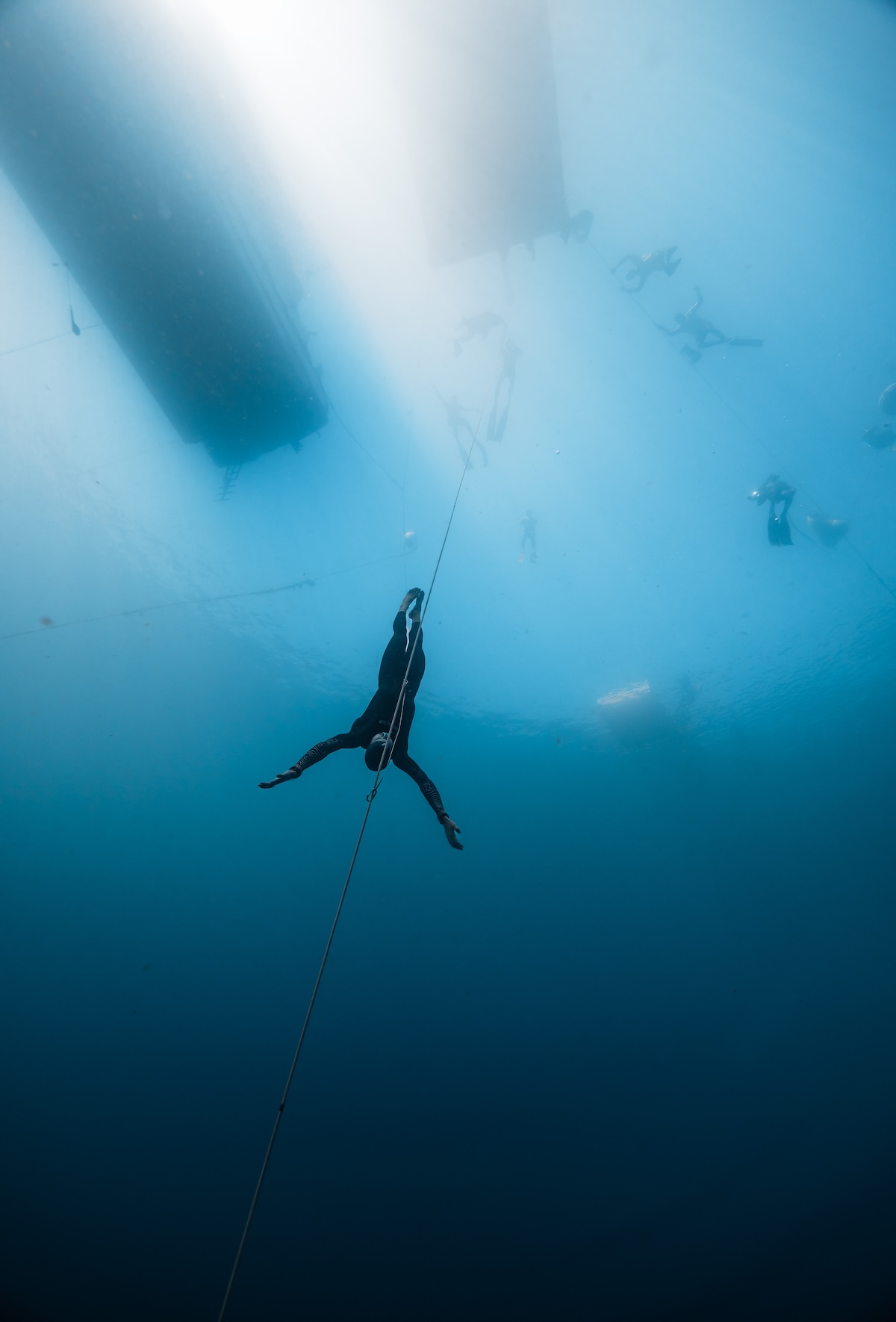 A freediver hanging motionless at depth beneath the platform, suspended in blue