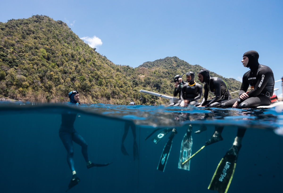 Freedivers at a Blue Element depth camp gathered at the surface platform, lush Dominica coastline behind, fins below the waterline