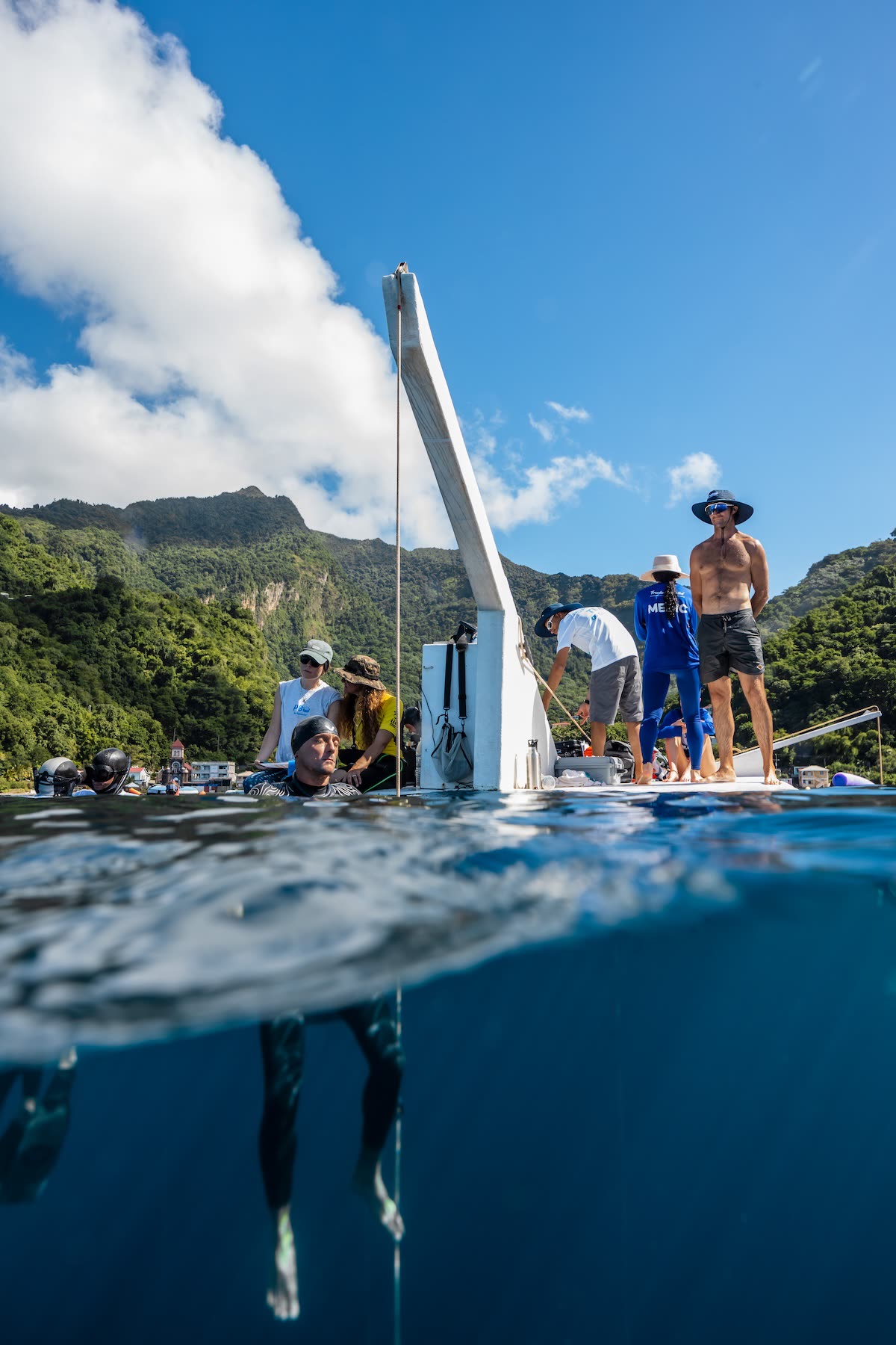 Over-under view at a competition platform in Soufrière Bay — the safety team and divers on the deck above the waterline, a freediver suspended on the line below, the green ridge of Dominica behind