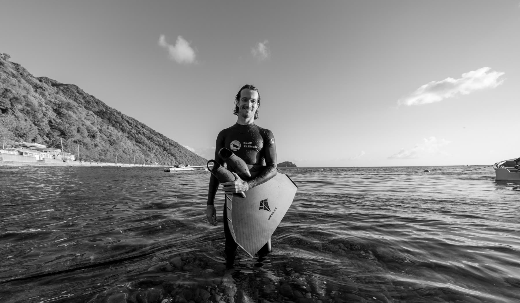 Harry McCahill standing in the shallows of Soufrière Bay in a Blue Element wetsuit, holding his fins, the forested flank of the bay rising behind him