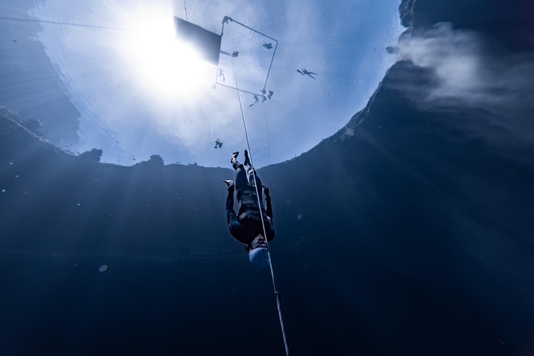 A competitor descending headfirst on the line into deep blue water, safety divers and the surface platform silhouetted against the sun above — Vertical Blue, where Harry McCahill was live commentator in 2023