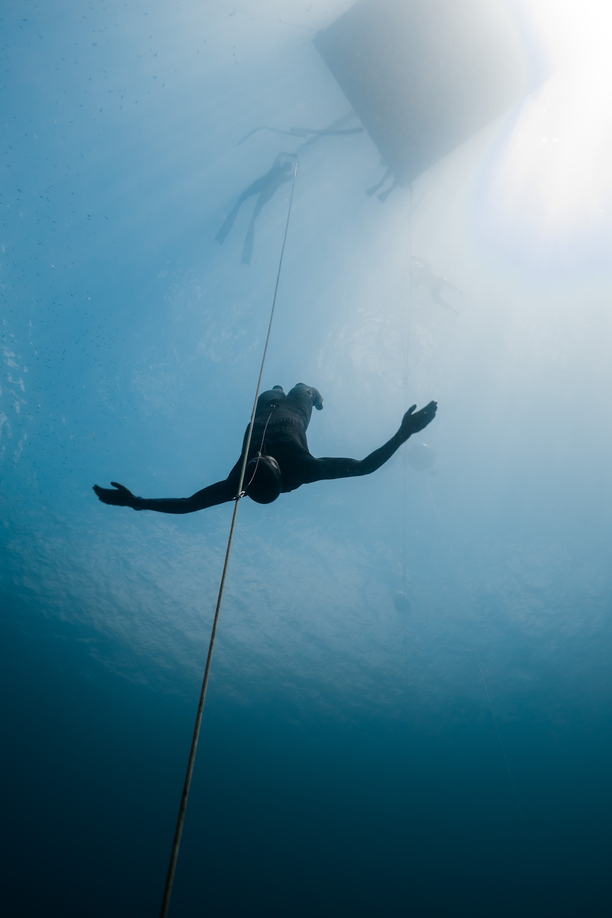 A no-fins freediver gliding weightless, arms spread, in a column of light from the surface